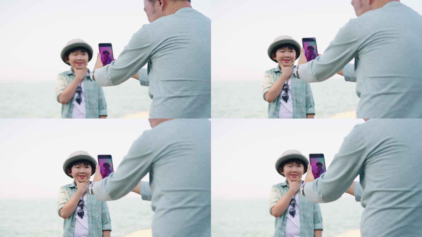 微笑的亚洲男孩扮演模特的特写镜头，他的父亲用智能手机拍照，一起在海滩上度过美好时光，背影