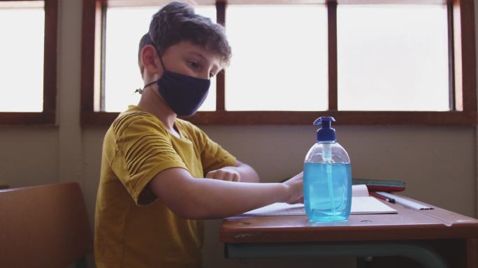 一个男孩坐在学校的课桌上，戴着口罩消毒双手