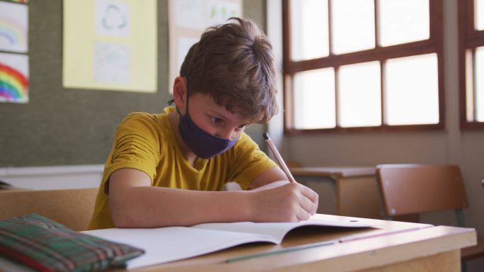 男孩戴着口罩在学校的桌子上写字