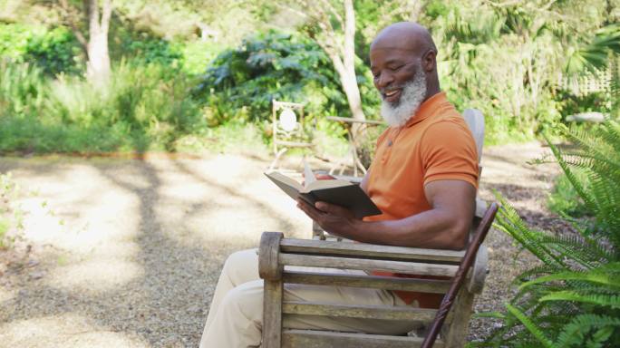 一位非裔美国老人微笑着坐在花园里的长凳上看书