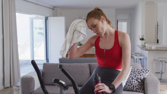 女人喝着水，坐在家里的健身自行车上用毛巾擦脸