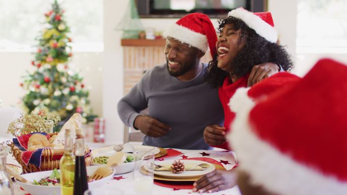 戴着圣诞老人帽的非裔美国夫妇微笑着坐在餐桌上敬酒吃午餐