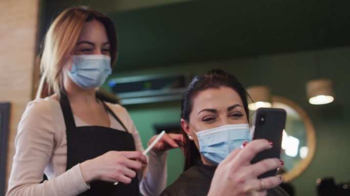 女发型师和女顾客戴着口罩在发廊看智能手机