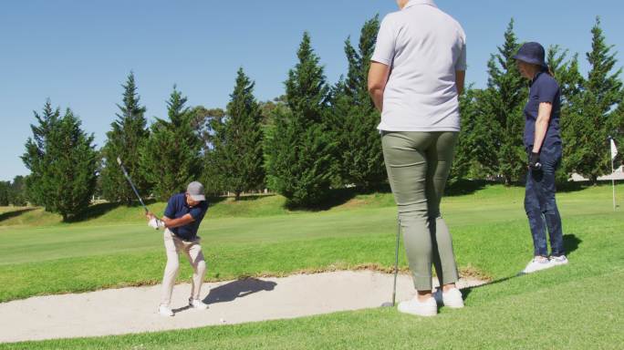 在一个阳光明媚的日子里，一个白人老人在高尔夫球场上从沙坑里打高尔夫球