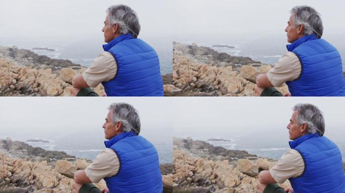 一位资深的徒步旅行者坐在山上的岩石上，看着海岸。