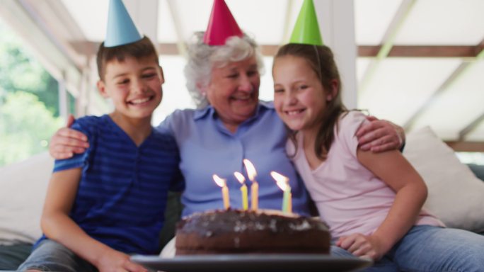 白人年长妇女在派对帽子拥抱她的孙子和生日蛋糕在桌上在家