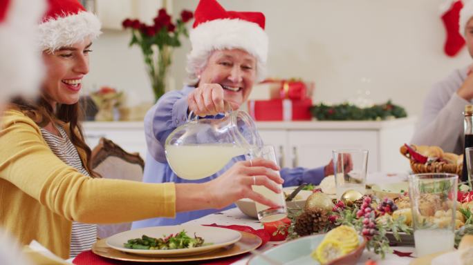 白种老妇人在圣诞老人的帽子倒饮料在玻璃杯中的女人，而坐在餐桌上一个