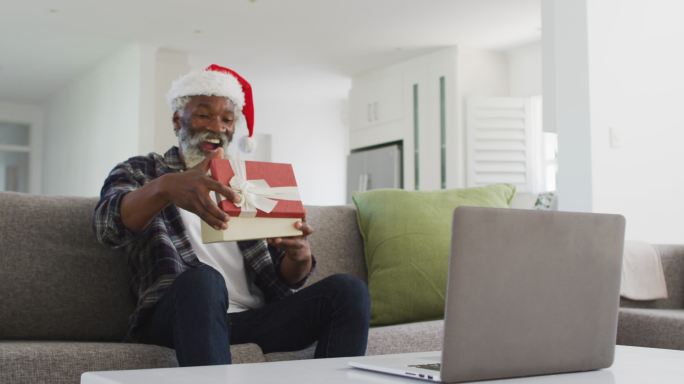 老人戴着圣诞老人帽打开礼物盒，同时在他的笔记本电脑上视频聊天