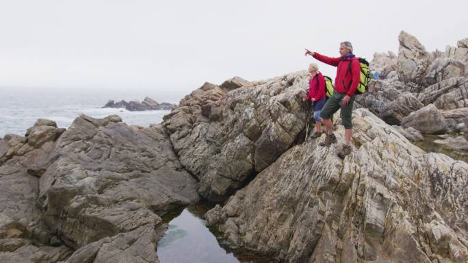 一对年长的徒步者夫妇站在岩石上，背着背包指向一个方向