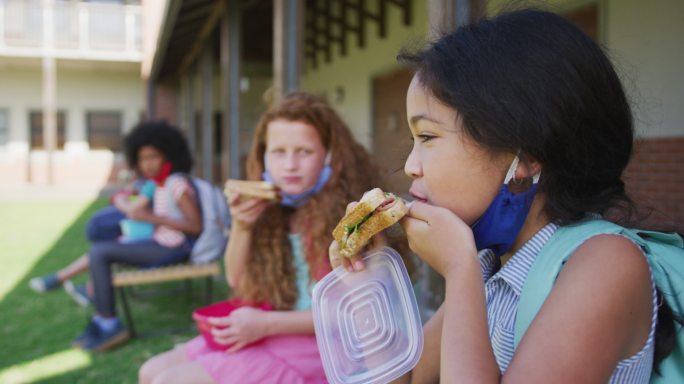 两个女孩坐在学校公园的长凳上吃午餐盒饭
