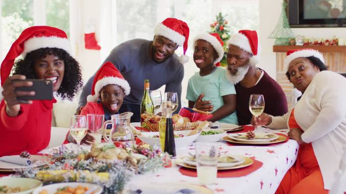 非裔美国家庭戴着圣诞老人帽坐在餐桌上用智能手机自拍哈哈