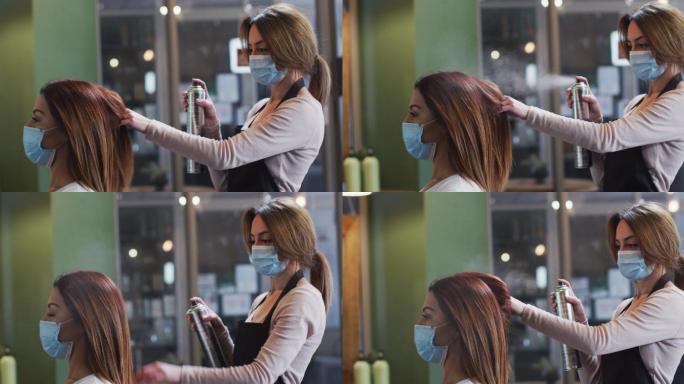 女发型师戴着面罩在发廊里向女顾客的头发喷发胶