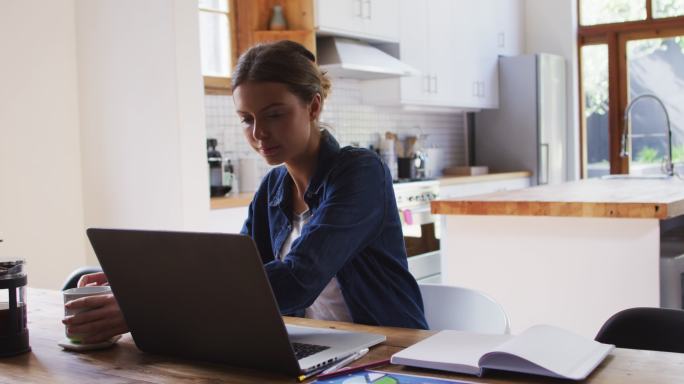 拿着咖啡杯的女人在厨房用笔记本电脑