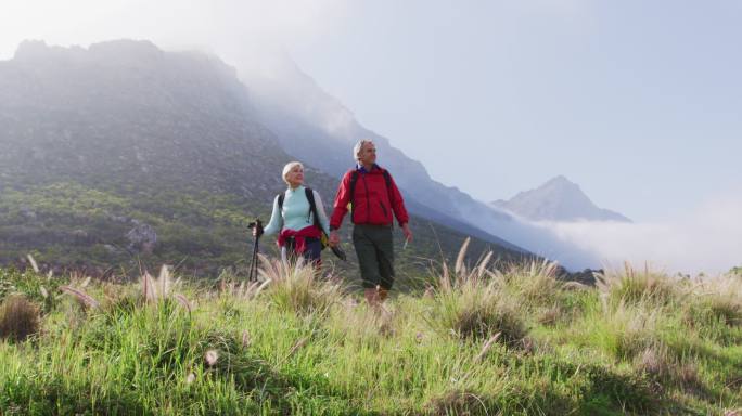 年老的徒步夫妇背着背包和登山杆，在徒步旅行时手牵着手走