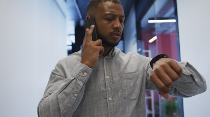 混血商人在现代化的办公室里一边用智能手机说话一边看手表
