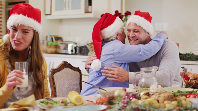 白种老妇人在圣诞老人的帽子拥抱她的儿子，而坐在餐桌上享受午餐