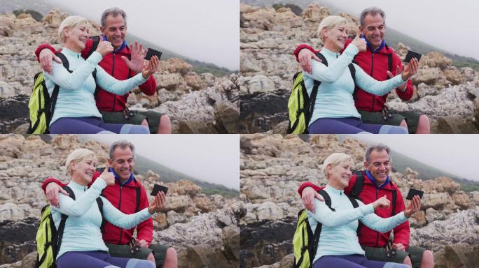 一对背着背包的老年徒步夫妇坐在岩石上，一边挥手一边进行视频通话