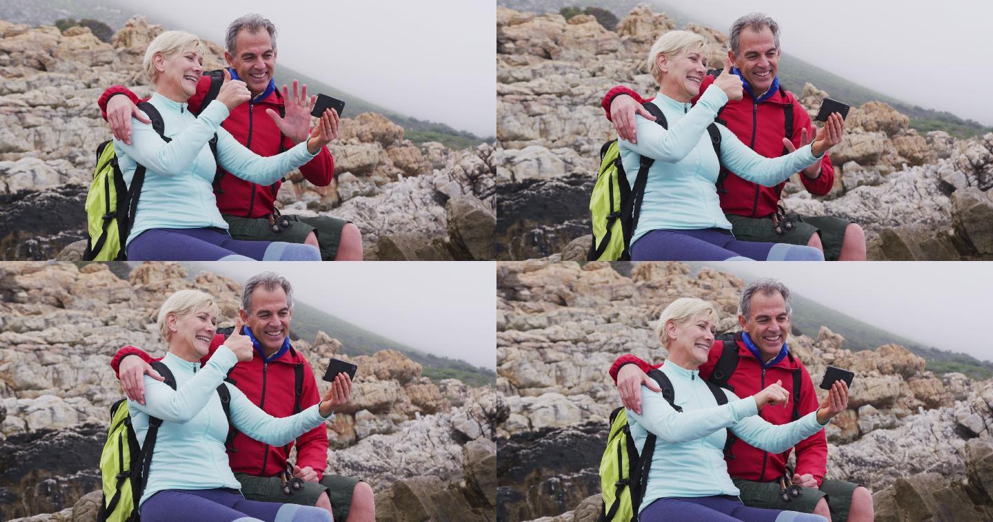 一对背着背包的老年徒步夫妇坐在岩石上，一边挥手一边进行视频通话