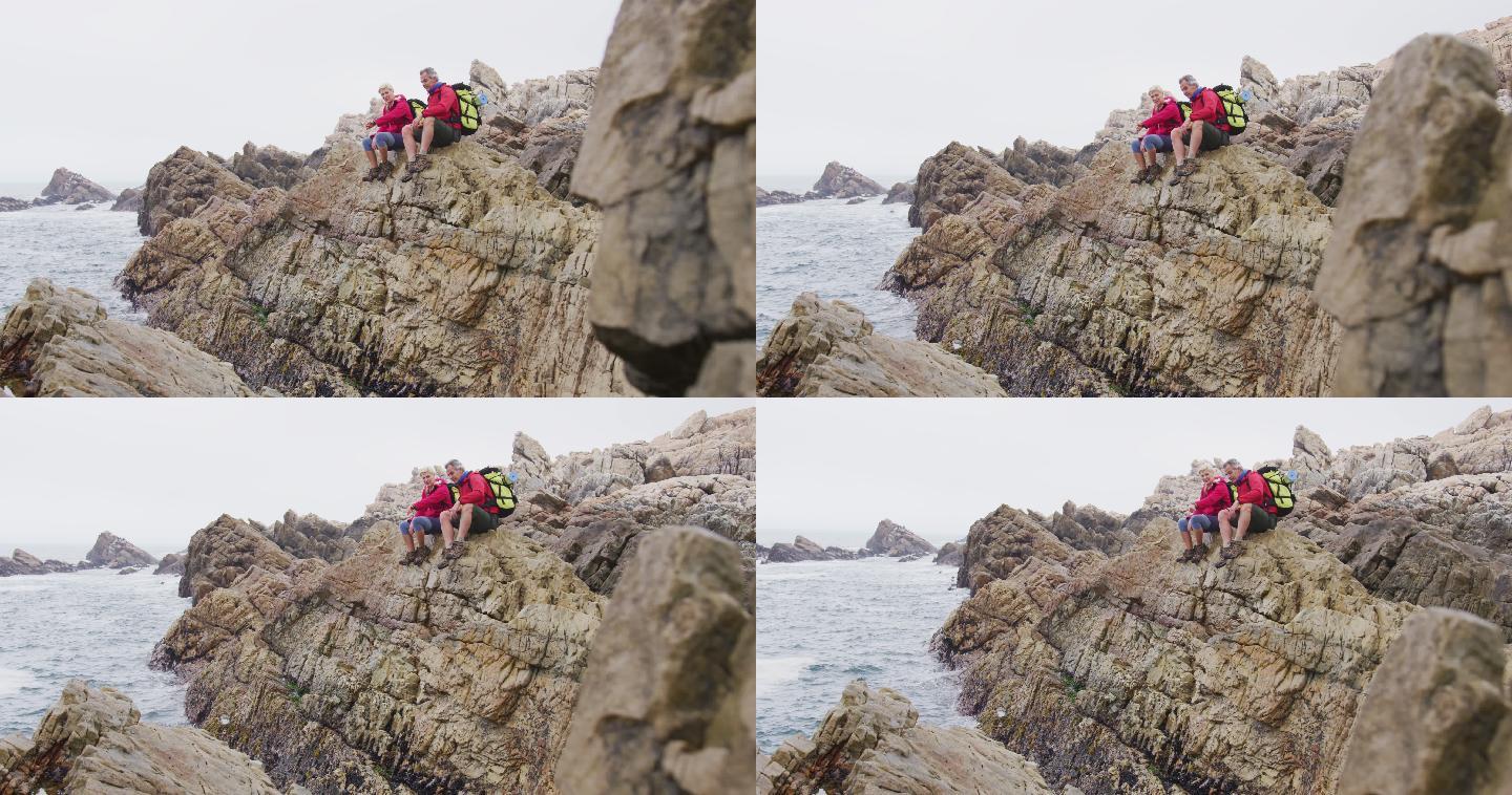 一对背着背包的老年徒步夫妇在海边徒步时坐在岩石上聊天。