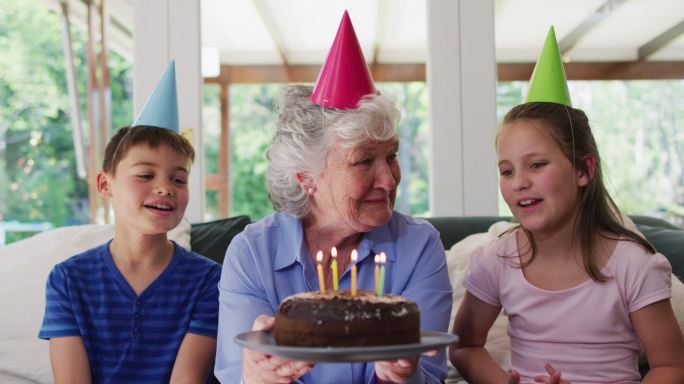 白种老妇人在派对帽子吹蜡烛在生日蛋糕，而她的孙子坐在