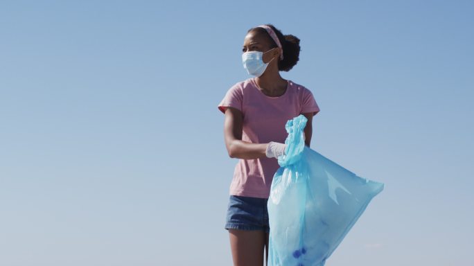 非洲裔美国妇女戴着口罩，拿着垃圾袋，在海滩上捡垃圾