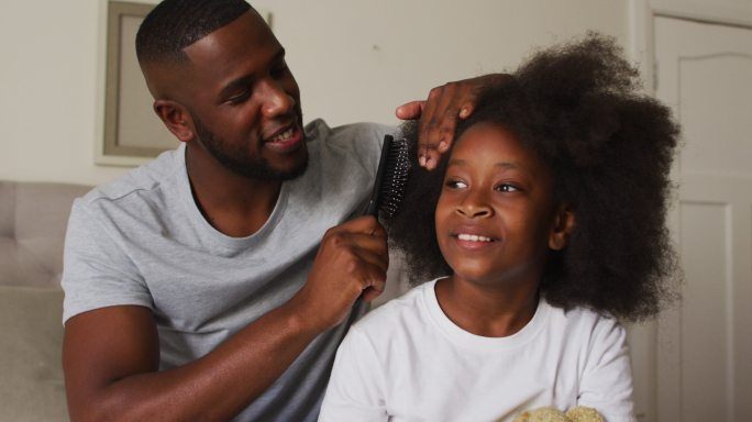 一位非裔美国父亲坐在家里的床上为女儿梳头