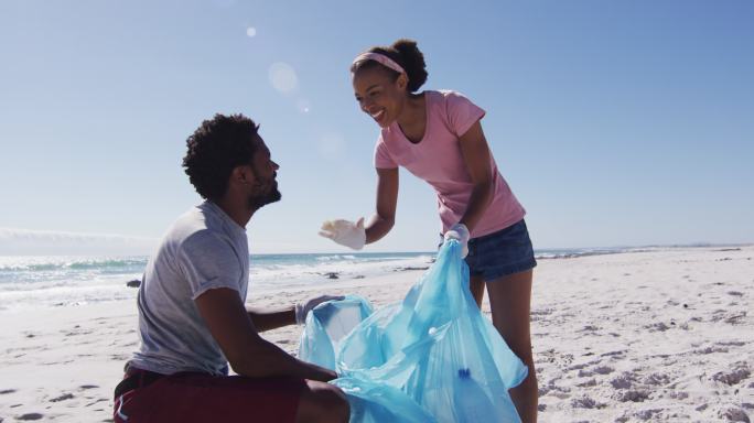 一对非洲裔美国夫妇举着垃圾袋，在海滩上捡垃圾，击掌庆祝