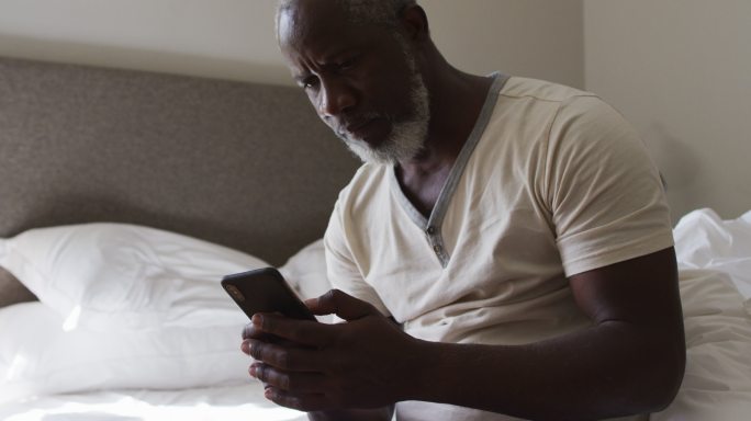 非裔美国老人坐在家里的床上使用智能手机