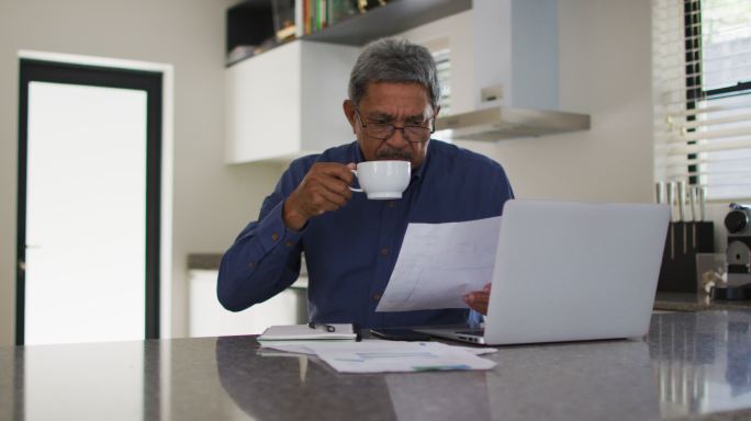 高级混合种族男子使用笔记本电脑和喝咖啡在厨房
