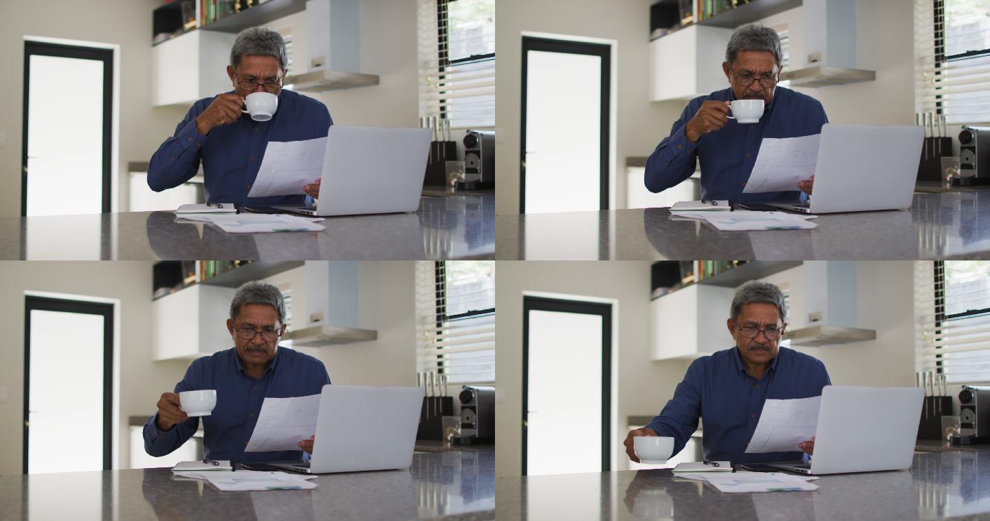 高级混合种族男子使用笔记本电脑和喝咖啡在厨房