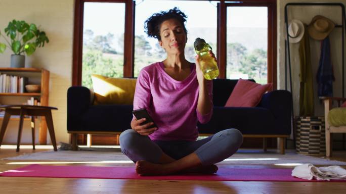 一名混血儿女子坐在瑜伽垫上一边喝水一边使用智能手机