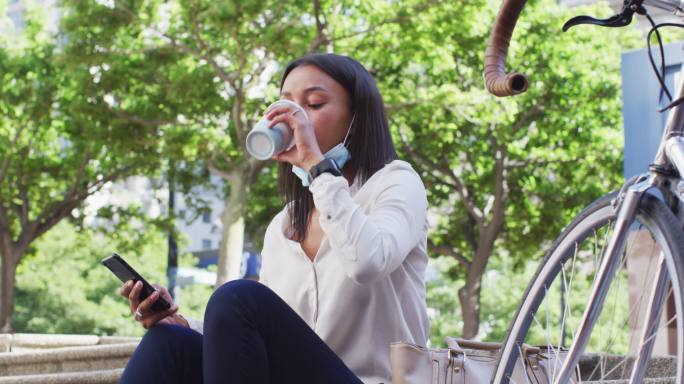 非裔美国妇女戴着口罩用智能手机在城市公园喝咖啡