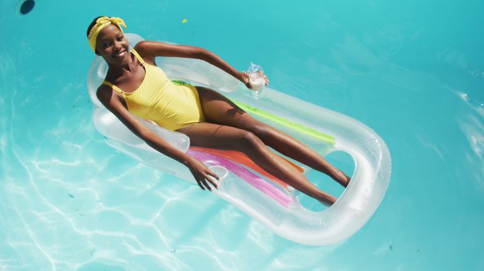 迷人的非洲裔美国女人躺在充气抱着饮料和微笑在游泳池
