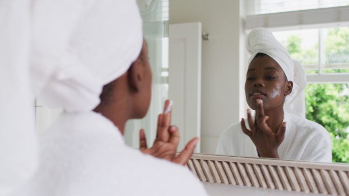 非裔美国妇女穿着浴袍，在家里照镜子时涂面霜