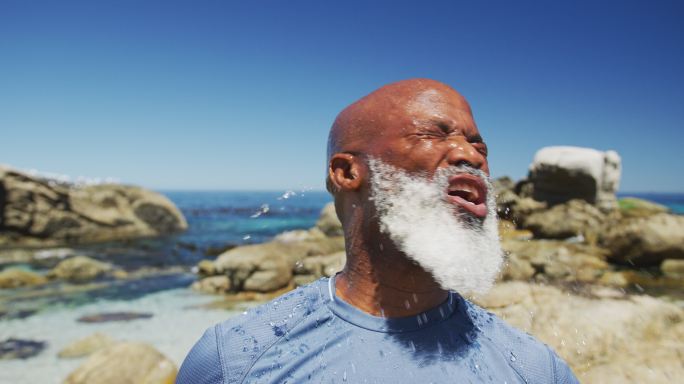 一位年长的非洲裔美国人在海边的岩石上练习往脸上浇水