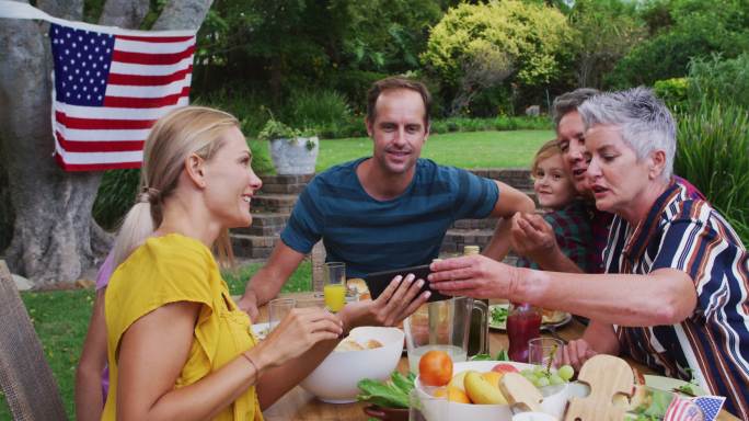 微笑的白人家庭庆祝餐在花园里一起看智能手机