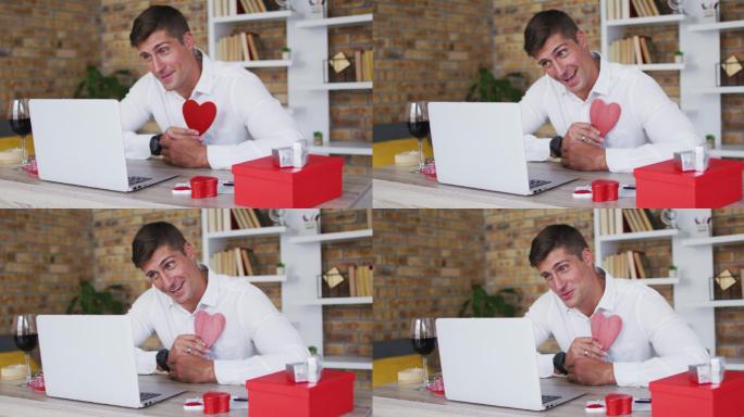 一名白人男子正在用笔记本电脑进行情人节视频通话，手持红心，面带微笑