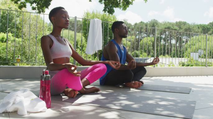 一对非洲裔美国夫妇练习瑜伽，坐在阳光灿烂的花园露台上沉思