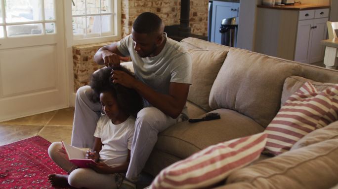 一位非裔美国父亲坐在家里的沙发上为女儿扎头发