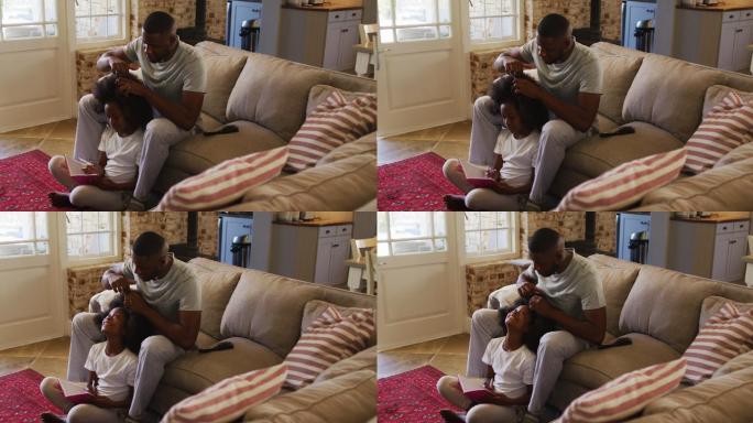 一位非裔美国父亲坐在家里的沙发上为女儿扎头发