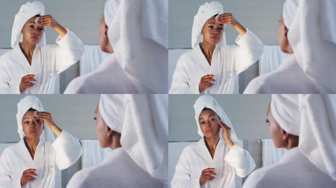非裔美国妇女在浴衣清洁她的脸与棉垫在浴室看着镜子