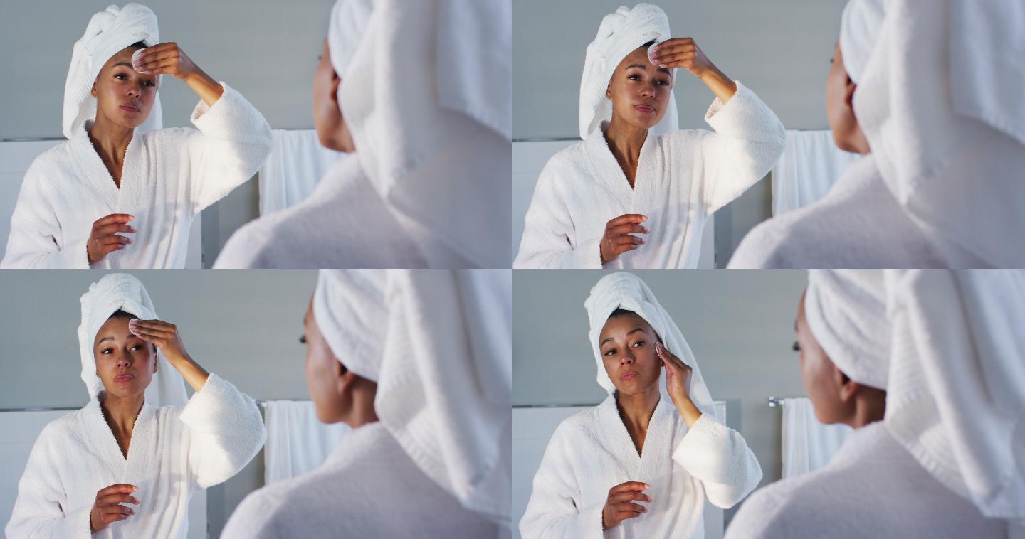 非裔美国妇女在浴衣清洁她的脸与棉垫在浴室看着镜子