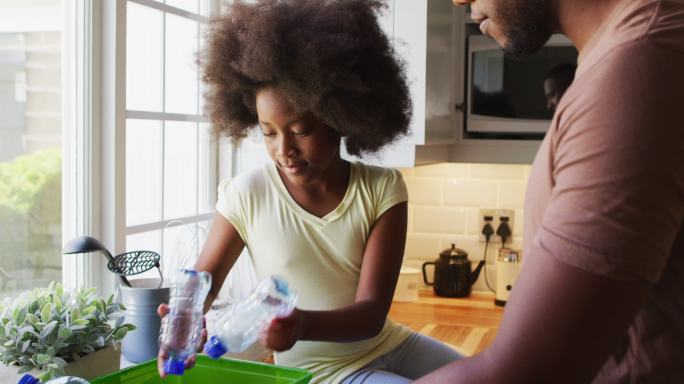 非洲裔美国人的女儿和父亲在厨房里整理回收品时玩塑料瓶