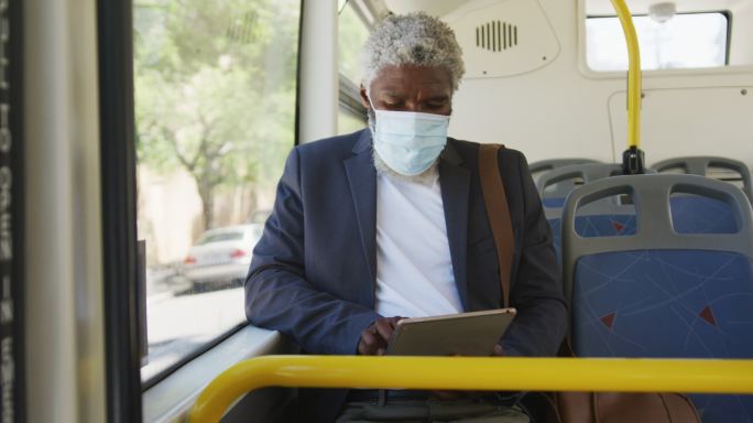 非裔美国老人戴着口罩坐在公交车上使用电子平板电脑