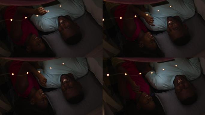 一个非裔美国人的女儿和她的父亲躺在有仙女灯的毯子帐篷里看平板电脑