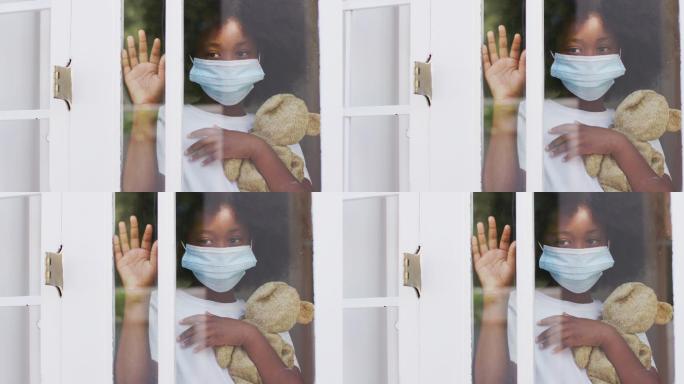 一个非裔美国女孩戴着面具，拿着泰迪熊在窗口招手