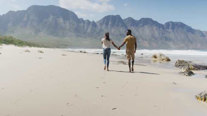 后视图的非洲裔美国夫妇牵着手，走在海滩上