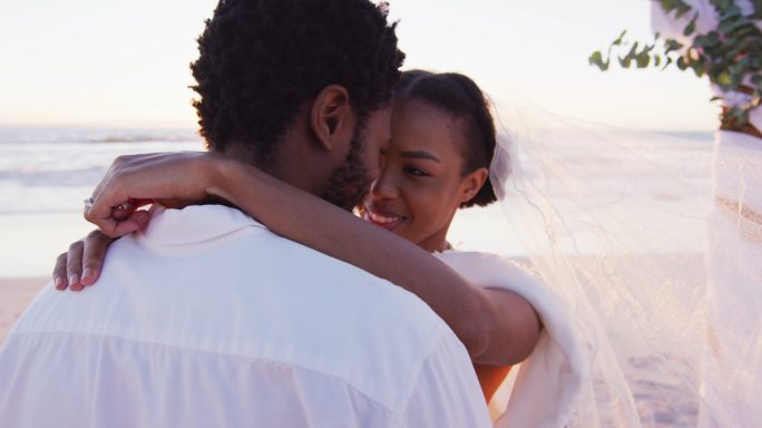 非洲裔美国人相爱的情侣结婚了，在日落的海滩上微笑着看着别人