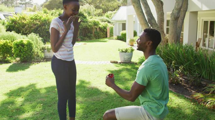 幸福的非裔美国夫妇在阳光灿烂的花园男人跪着拿着戒指向女人求婚