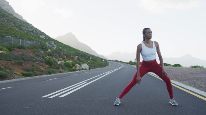 适合混合种族妇女锻炼伸展在一个乡村公路附近的山